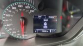Car dashboard showing speedometer up to 140 mph, digital readout at 0 mph with trip and vehicle screens, and 76,590 miles on the odometer; seatbelt warning lit.