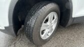 Close-up of a light-colored car wheel and tire, with a wet tread pattern and rain droplets on the body and pavement below.
