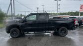Black pickup truck parked in a wet parking lot, with power lines and a cloudy sky overhead and other cars in the background.