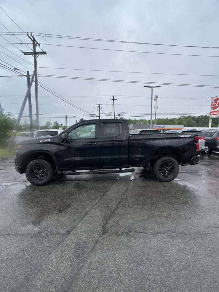 Black pickup truck parked in a wet parking lot, with power lines and a cloudy sky overhead and other cars in the background.