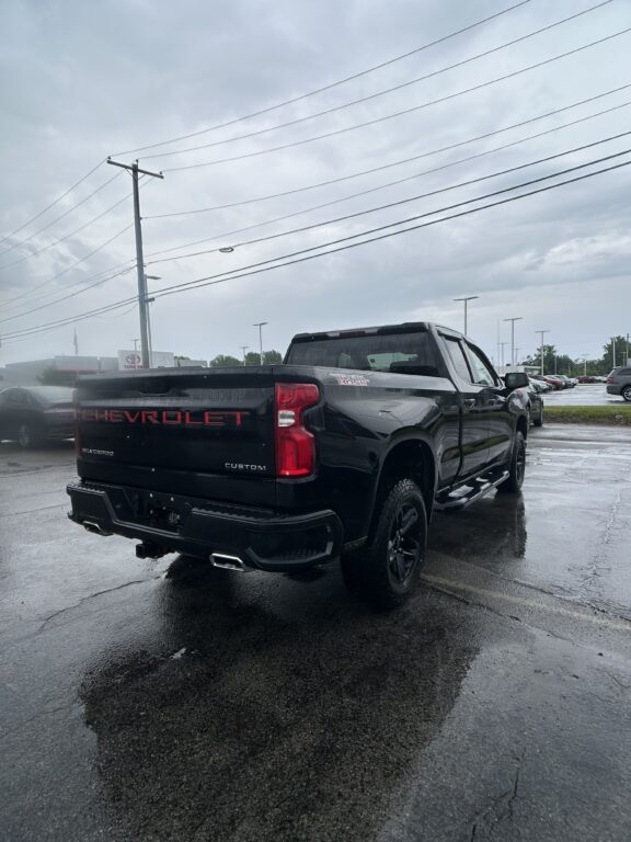Rear three-quarter view of a black Chevrolet Silverado pickup in a rainy dealership lot, with power lines overhead and wet pavement.