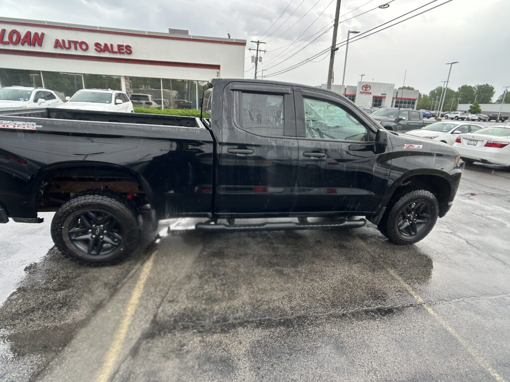 Black pickup truck parked in a dealership lot with a white building labeled 'AUTO SALES' in the background and other cars nearby on a wet pavement.
