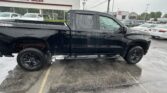 Black pickup truck parked in a dealership lot with a white building labeled 'AUTO SALES' in the background and other cars nearby on a wet pavement.