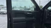Driver-side door of a black pickup truck open, raindrops on the window, parking lot with cars visible outside.