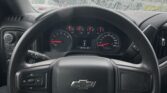 Driver's view of a car interior with a rain-speckled windshield; dashboard gauges visible behind a black steering wheel bearing a Chevrolet emblem on the airbag cover.