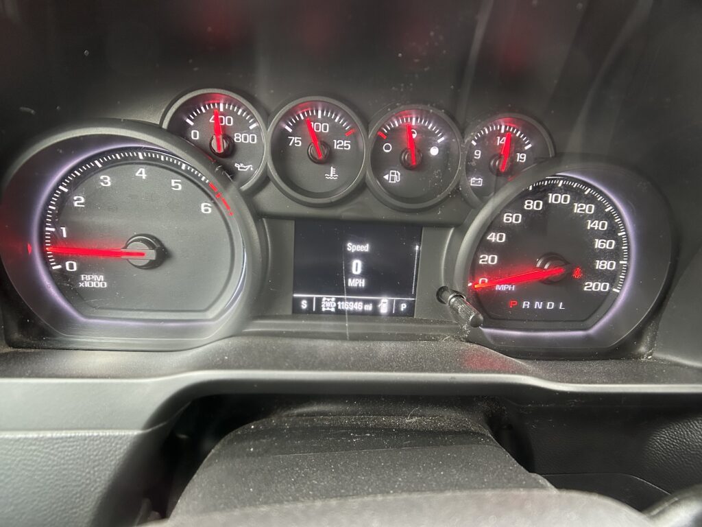 Close-up of a car dashboard showing circular gauges with red needles and a central screen displaying Speed 0 MPH (P R N D L gear indicator).