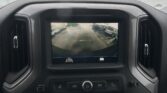Dashboard screen showing a rearview camera view of a parking lot with parked cars, as rain beads cling to the windshield outside.