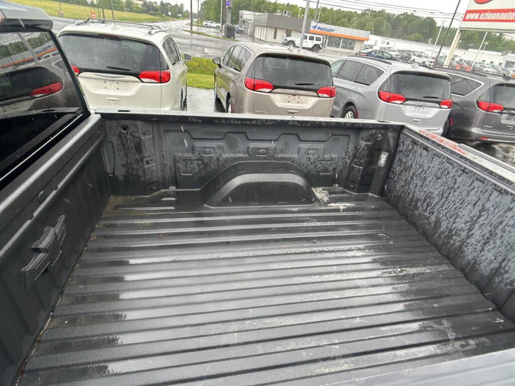 View from inside a pickup truck bed looking toward a row of silver and gray minivans parked in a car lot on a rainy day.
