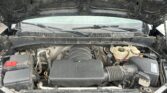 Open car hood showing engine bay with battery, air intake, and coolant reservoir; dusty, with wiper blades visible in the windshield area beyond.
