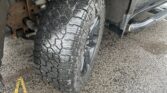 Front wheel and large, rugged tire of a black pickup truck, with visible suspension and rust, on a wet paved surface with yellow lines nearby.