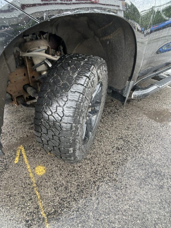 Front wheel and large, rugged tire of a black pickup truck, with visible suspension and rust, on a wet paved surface with yellow lines nearby.