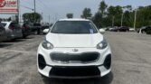 Front view of a white Kia compact SUV parked in a dealership lot with other cars and a blue sky above.