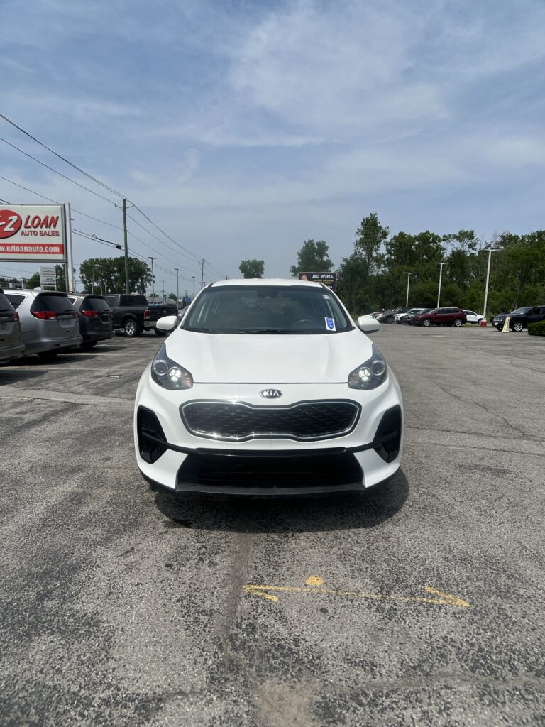 Front view of a white Kia compact SUV parked in a dealership lot with other cars and a blue sky above.
