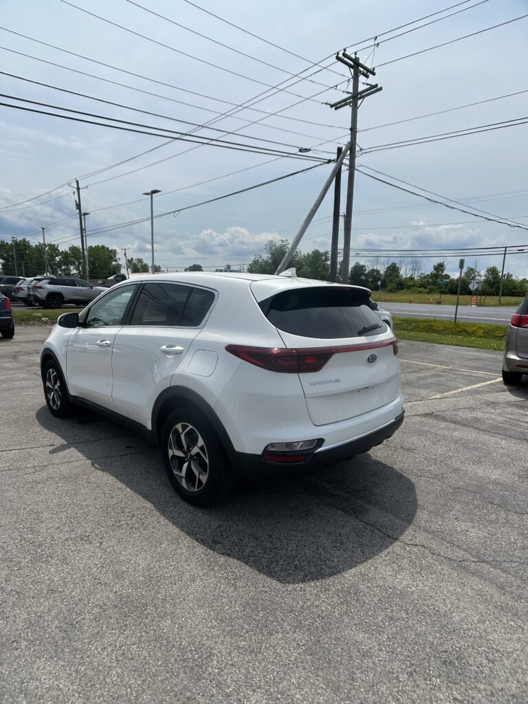 White Kia Sportage SUV parked in a lot with utility poles and power lines overhead, blue sky in the background.