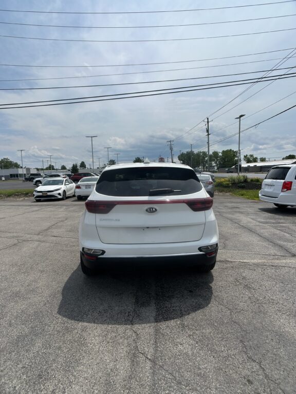 Rear view of a white Kia SUV in a parking lot, with several other cars and tall power lines overhead under a blue sky.