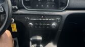 Inside view of a car dashboard with a central touchscreen showing radio station 98.1 and car icons, gear shifter in front, and cup holders nearby