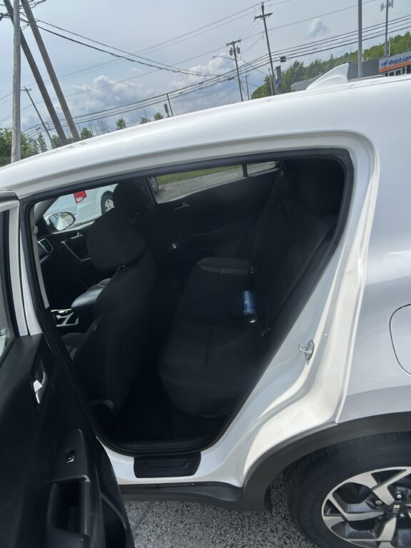 Open rear passenger door of a white SUV, showing black fabric seats and a blue water bottle on the back seat, with a parking lot outside.