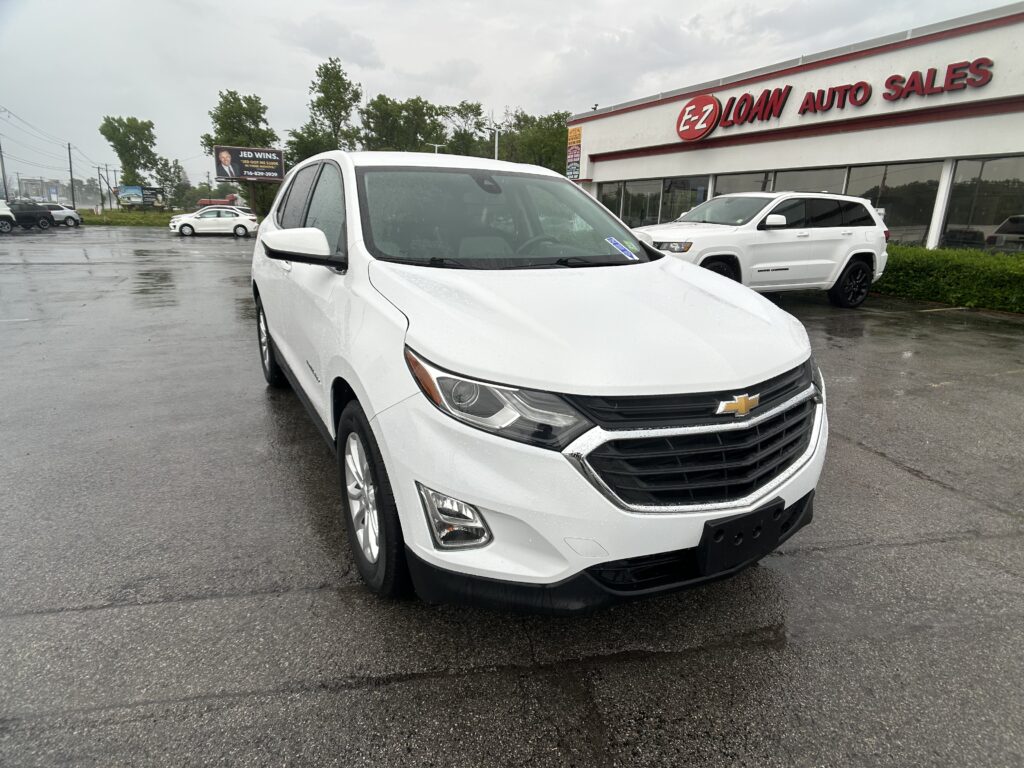 White Chevrolet SUV in a car lot with EZ Loan Auto Sales building signage in the background on a rainy day, reflections on wet pavement.