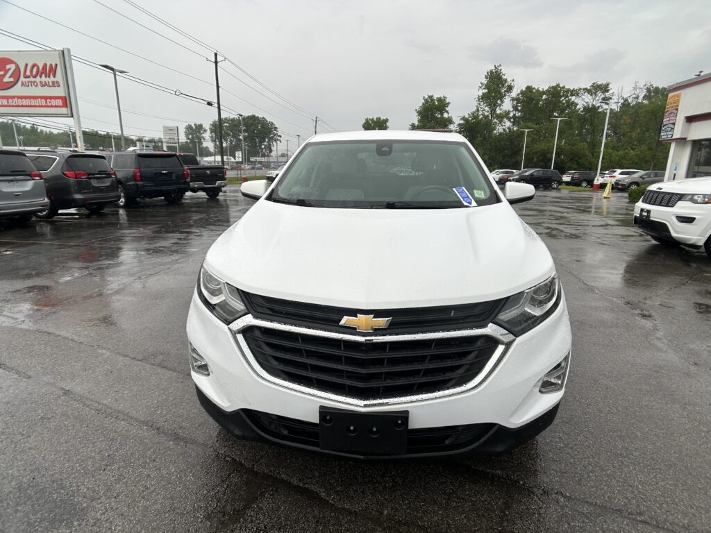 Front view of a white Chevrolet SUV in a car dealership lot on a rainy day, with wet pavement and other vehicles in the background.