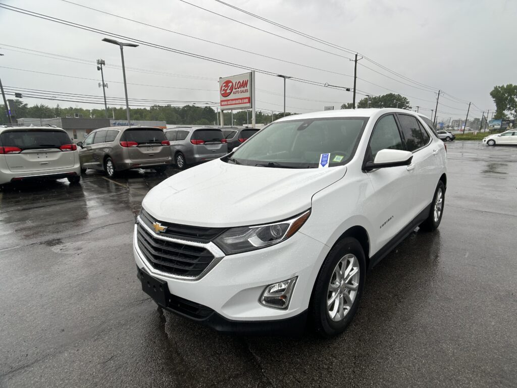 Front-left view of a white Chevrolet SUV in a rainy car dealership lot, with a row of minivans behind and an EZ LOAN Auto Sales sign in the background.