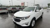 Front-left view of a white Chevrolet SUV in a rainy car dealership lot, with a row of minivans behind and an EZ LOAN Auto Sales sign in the background.