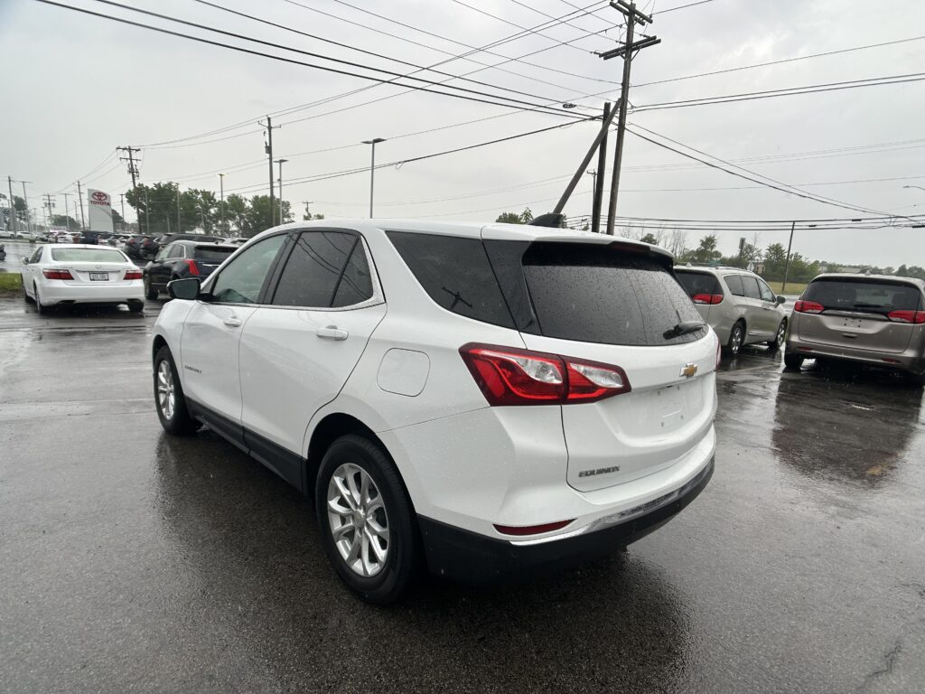 White Chevrolet Equinox in the foreground of a rainy car dealership lot, with many cars, wet pavement, and overhead power lines.