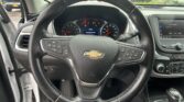 Steering wheel with Chevrolet emblem in foreground, instrument cluster and infotainment screen visible, parking lot outside the windshield.