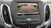 Rearview camera display showing a wet parking lot behind the car, with parked vehicles and a building in the background, inside the vehicle dashboard panel.