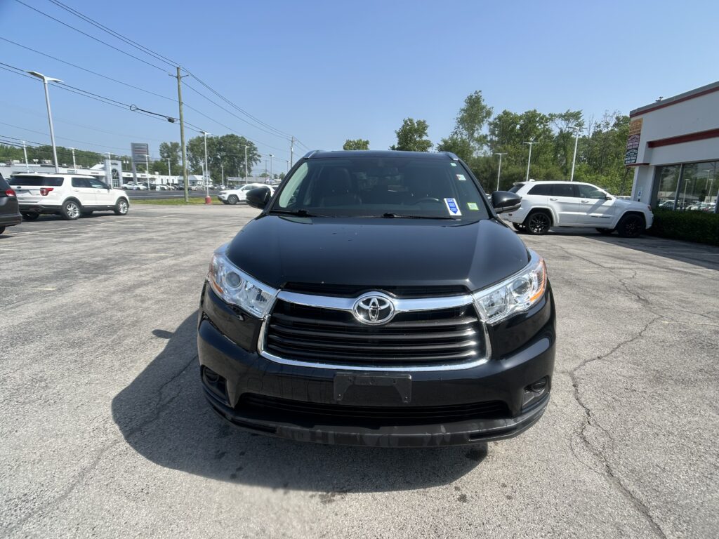 Front view of a black Toyota SUV in a parking lot near a dealership, with other cars and a white building on the right under a clear blue sky.