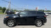 Close-up of a black SUV in a parking lot with utility poles and power lines, dealership sign and an American flag in the distance.