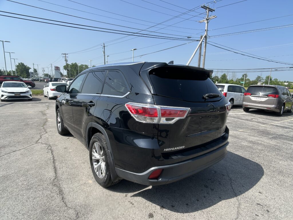 Rear three-quarter view of a black Toyota Highlander SUV in a busy dealership parking lot with other cars and overhead power lines against a clear blue sky.