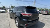 Rear three-quarter view of a black Toyota Highlander SUV in a busy dealership parking lot with other cars and overhead power lines against a clear blue sky.