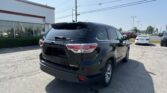 Black Toyota Highlander XLE AWD parked in a dealership lot with a white building on the left and rows of cars in the background under a clear sky.