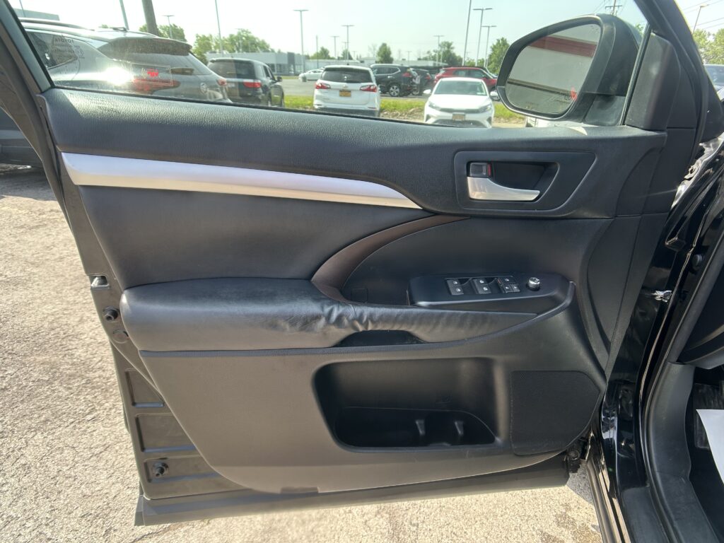 Driver-side car door interior panel with window controls and chrome handle; view of parking lot with multiple cars through the open window and side mirror outside