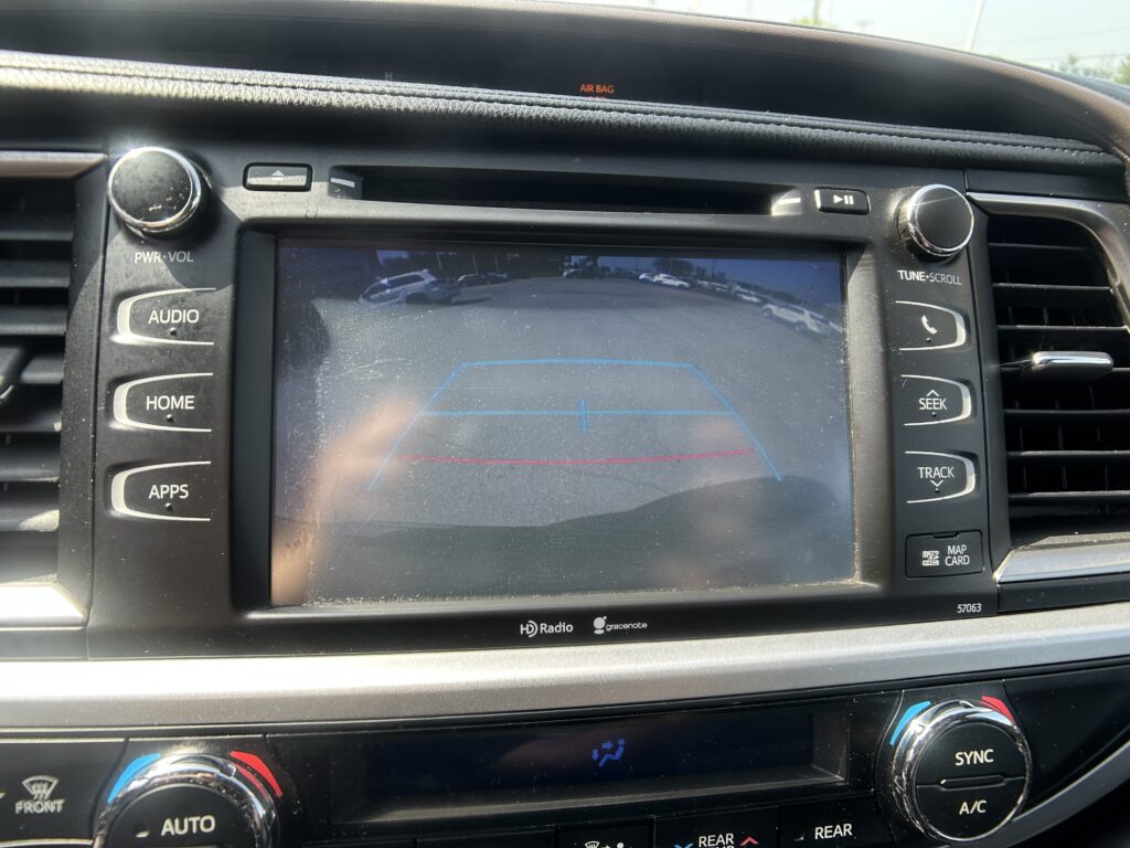 Center console of a car showing a touchscreen navigation/rear-view camera display with blue guide lines, flanked by physical audio controls and climate dials.