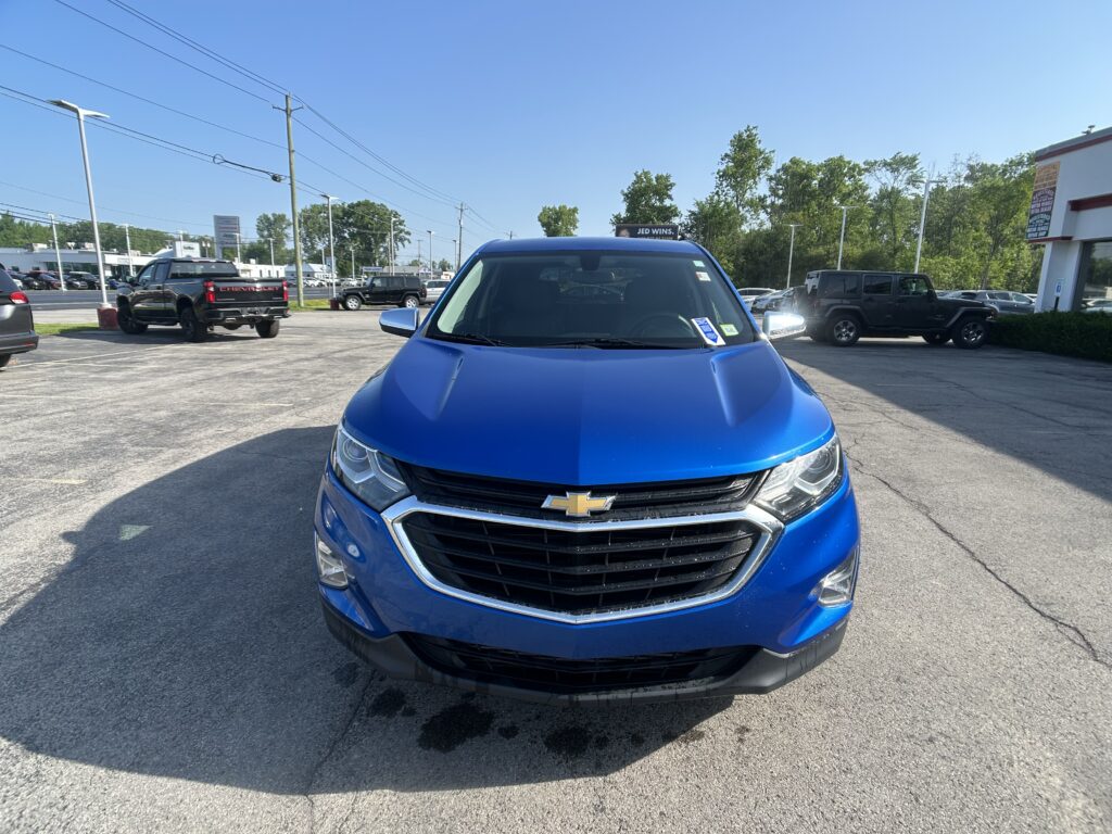 Front view of a blue Chevrolet SUV in a dealership parking lot, with other vehicles and a showroom building in the background.