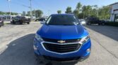 Front view of a blue Chevrolet SUV in a dealership parking lot, with other vehicles and a showroom building in the background.