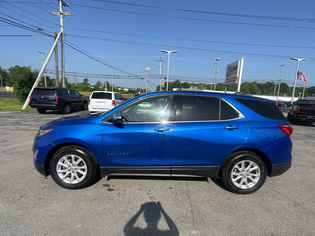 Blue Chevrolet Equinox SUV parked in a dealership lot with other cars, power lines, and an American flag in the background.