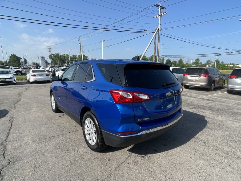 Blue Chevrolet Equinox in a dealership parking lot, with many other cars and overhead power lines under a clear blue sky, rear three-quarter view in foreground.