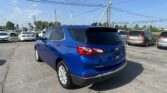 Blue Chevrolet Equinox in a dealership parking lot, with many other cars and overhead power lines under a clear blue sky, rear three-quarter view in foreground.