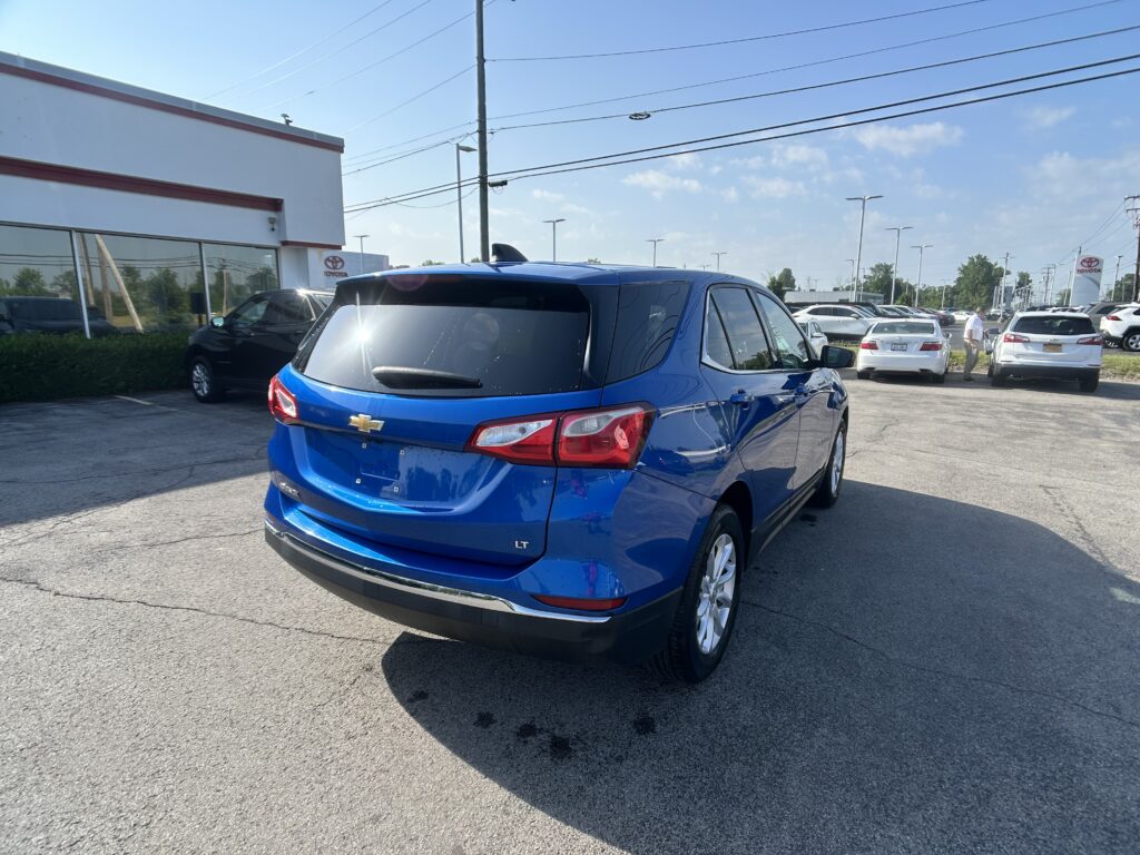 Blue Chevrolet SUV in a car dealership lot with a Toyota showroom and row of cars under a clear blue sky.