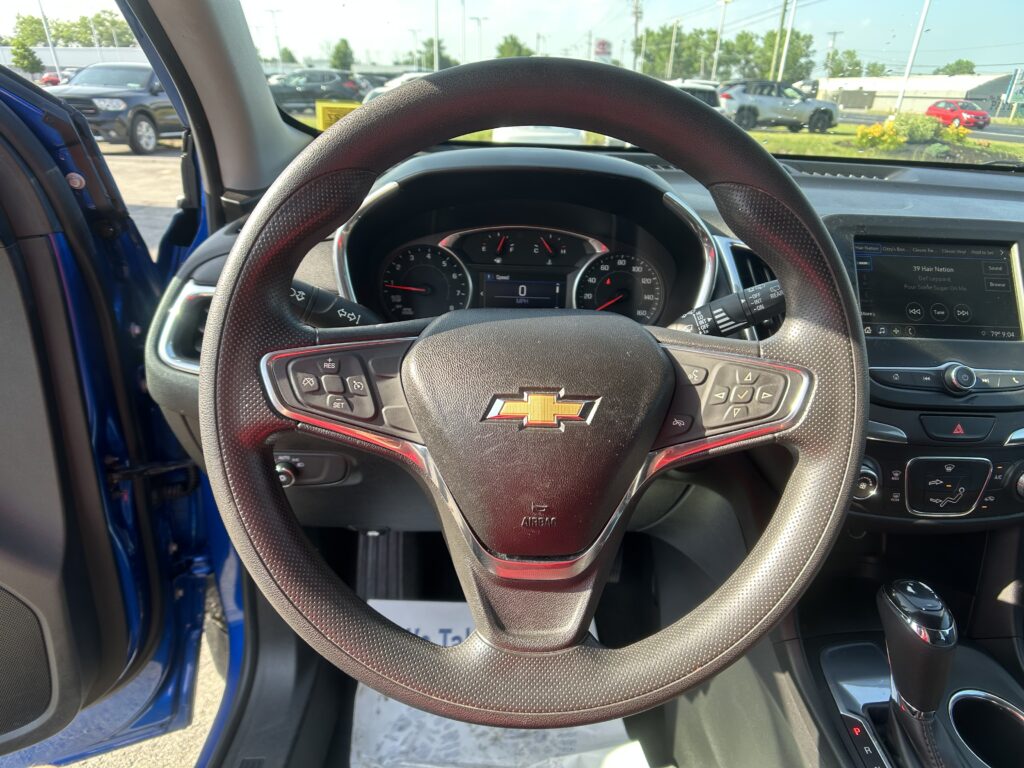 Driver's view of a Chevrolet interior: steering wheel, instrument cluster, and touchscreen with the driver's door open to a sunny parking lot outside.