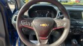 Driver's view of a Chevrolet interior: steering wheel, instrument cluster, and touchscreen with the driver's door open to a sunny parking lot outside.