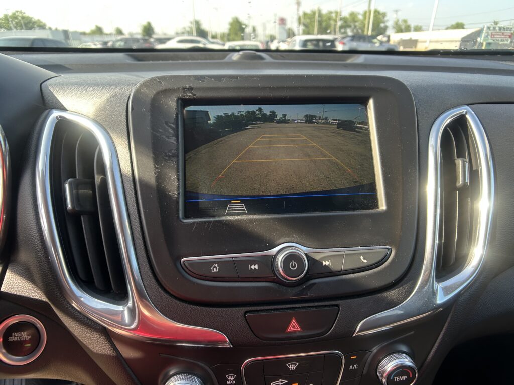In-dash rearview camera feed showing yellow guide lines over a parking lot, visible through the car's screen and console.