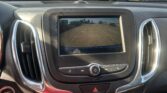 In-dash rearview camera feed showing yellow guide lines over a parking lot, visible through the car's screen and console.
