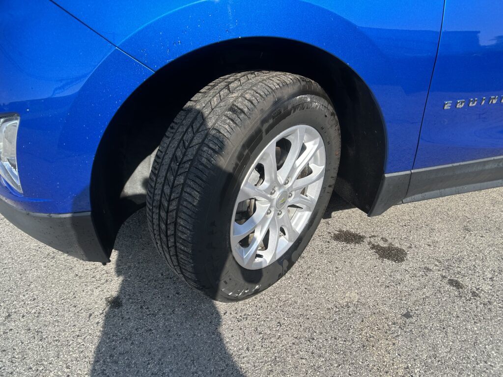 Close-up of a blue SUV wheel and tire, showing the front fender and wheel well area on a sunny day.