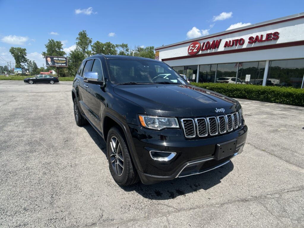 Black Jeep SUV parked diagonally in a lot outside an EZ Auto Sales dealership, with a blue sky and trees in the background.