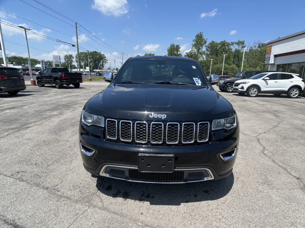 Front view of a black Jeep SUV in a car dealership lot with other vehicles and a building in the background.