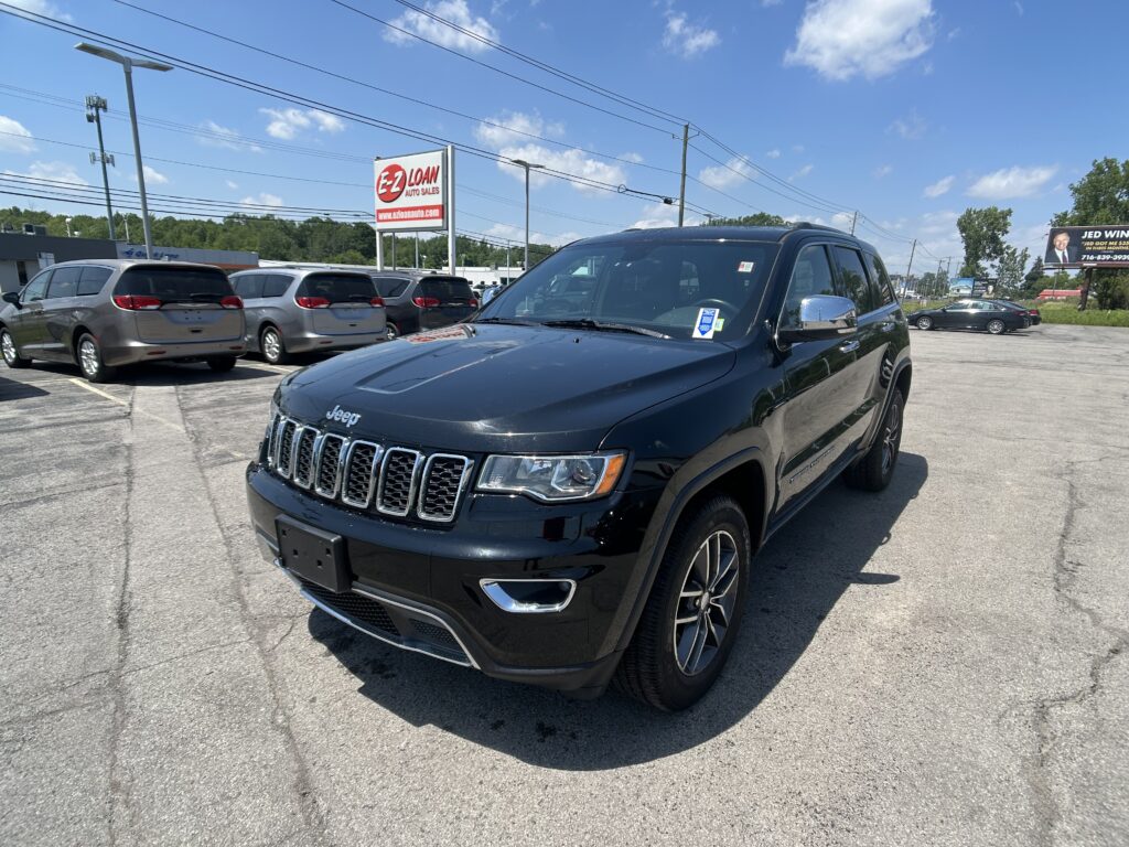 Close-up of a black Jeep SUV in a dealership parking lot with several minivans and an E-Z LOAN Auto Sales sign in the background on a sunny day.