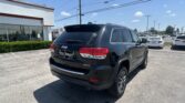 Rear view of a black Jeep Grand Cherokee Limited in a dealership lot on a sunny day, with a white building and power lines in the background.